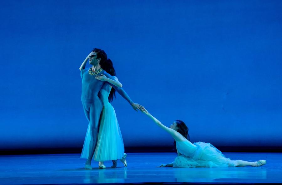 Three dancers are center stage. Two stand while one lies on the ground. The dancer on the ground has their arm extended, and fingertips touching the hand of the one of the standing dancers.