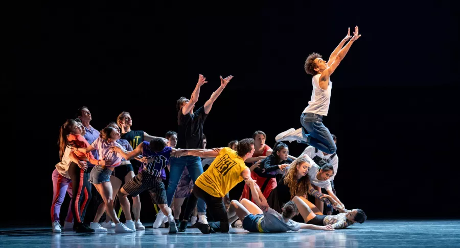 A large group of dancers, wearing an assortment of colors, range from standing to lying positions, all with their arms touching the person next to them. In the center, a dancers stands tall with their arms extended to the ceiling.