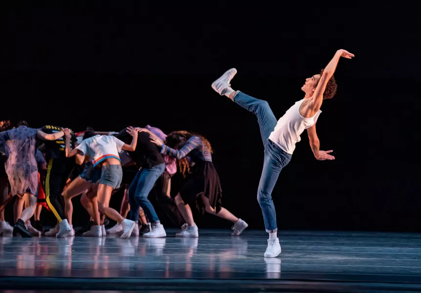 A dancer on stage right strikes a pose on one leg, with remaining arms and leg extended. On stage left a circular huddle of dancers are facing with their backs to the audience, with their hands all placed on each others backs.
