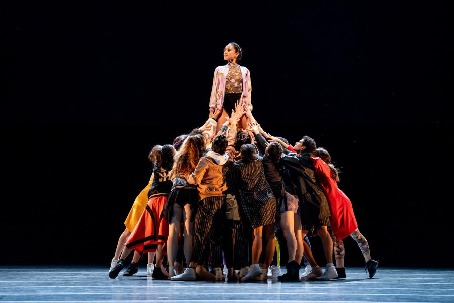 A large group of dancers wearing an assortment of reds, blacks and oranges, are huddled together, lifting a dancer above the group. The dancer being lifted poses with their arms to their side, wearing a light pink jacket.