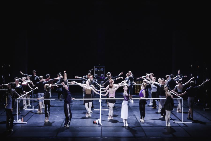 A large stage is outlined by a hollow square of connecting ballet barres. Dancers outline and interior and exterior of the square, all posed in fifth position.