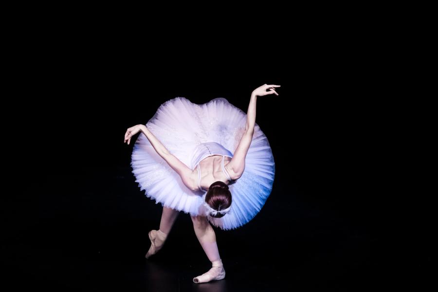 A ballerina wearing a white tutu, crown and pointe shoes, hits a low curtsy position, with their upper back facing the audience as they bend forward.