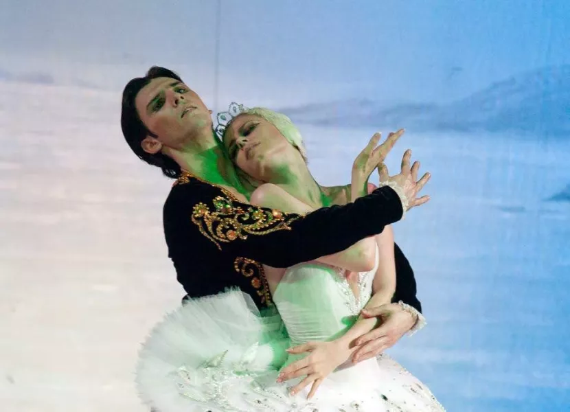 A ballerina wearing a white tutu and crown, is encapsed by a dancers with short dark hair, in a dark long sleeve velvet leotard with gold embellishments. The backdrop is a faded ombre of blue in the upper right, that turns to white in the bottom left.