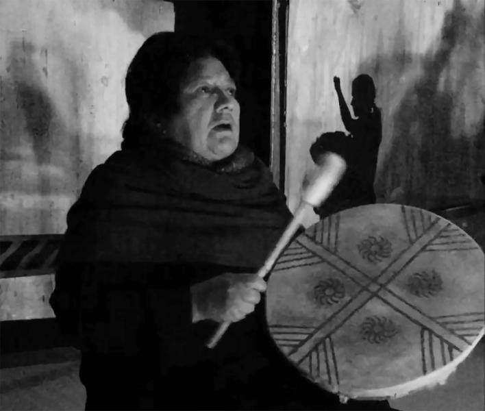 A black and white picture depicts a person wearing all black holding a drum and mallet. The drum has an X through it and each quarter has a design made up of straight lines toward the top and swirls near the middle. There is a shadow of another person hitting a drum in the background. 