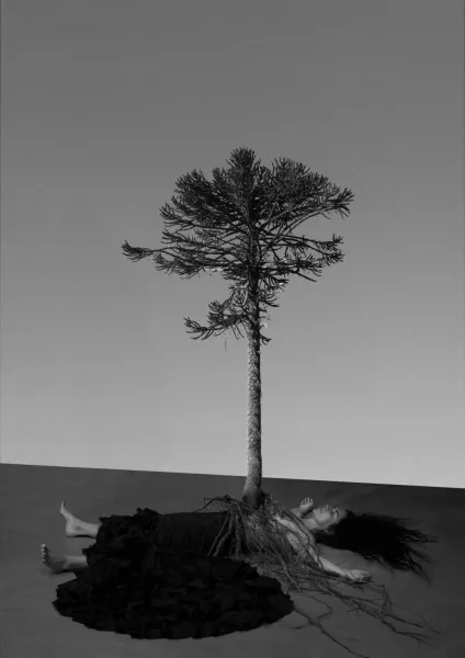 A black and white image depicts a person laying on the floor with their arms overhead. A tree's roots cover the person's body and the tree appears to be coming out of the person's torso. 