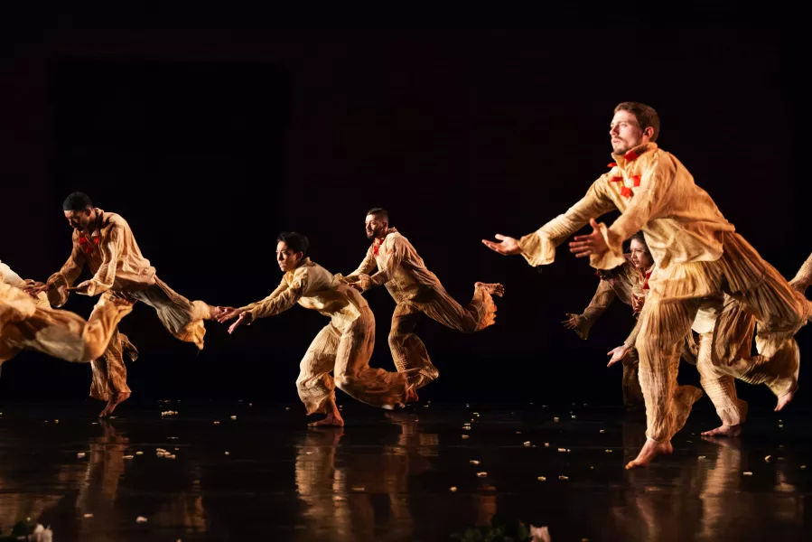 A group of dancers wearing matching tan shirts and pants, dance on an all black stage and backdrop, all striking the same pose: leaning forward with their arms both stretched before them.