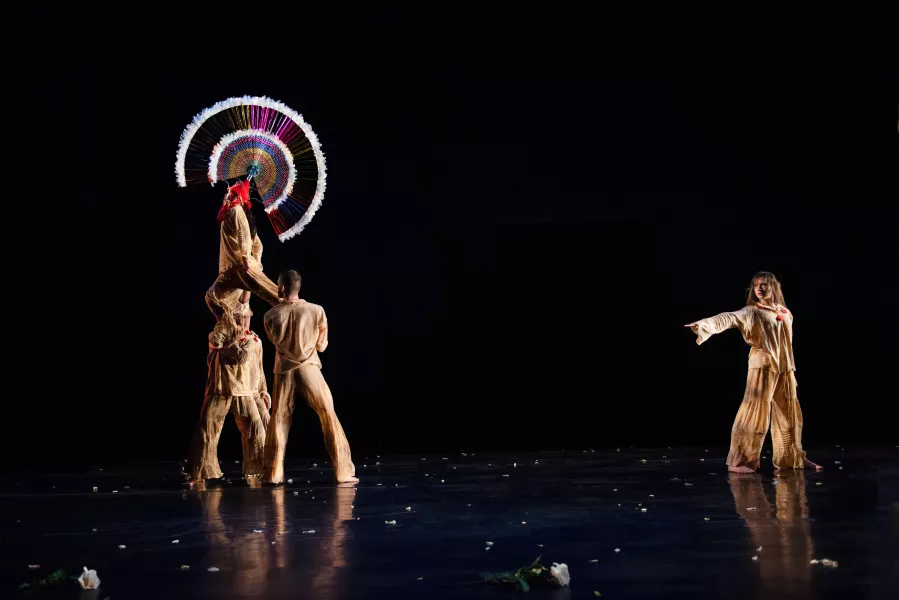 Two dancers hold a third above their shoulders, while a fourth dancers across the stage points a finger at the elevated dancer. The elevated dancer wears a large, circular headpiece, that stands out before the all black background. 