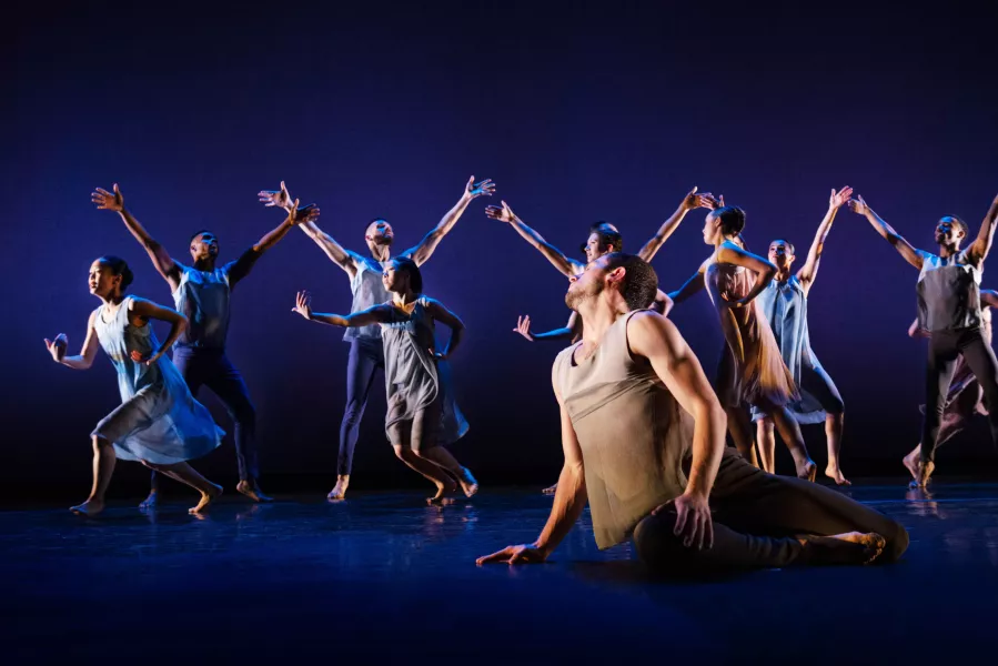 A dancer sits downstage with their head looking upstage. Upstage are two rows of dancers, the back with their arms stretched above their heads, the front with their arms stretched to their side.