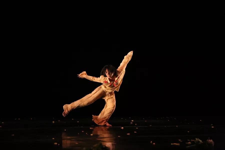 A dancer wearing a matching tan pants and shirt, strikes a standing pose on one leg, with their other leg extended to the side, and both arms above their head. The backdrop and floor beneath are solid black.
