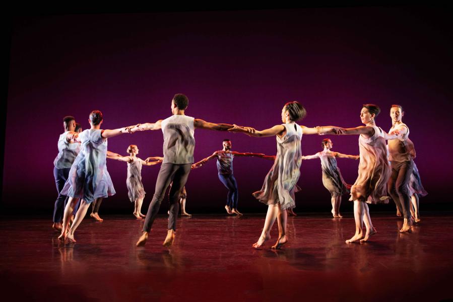 A group of dancers formed in a circle all holding each other by the wrists. The stage is lit with an ombre of black, purple and pink.