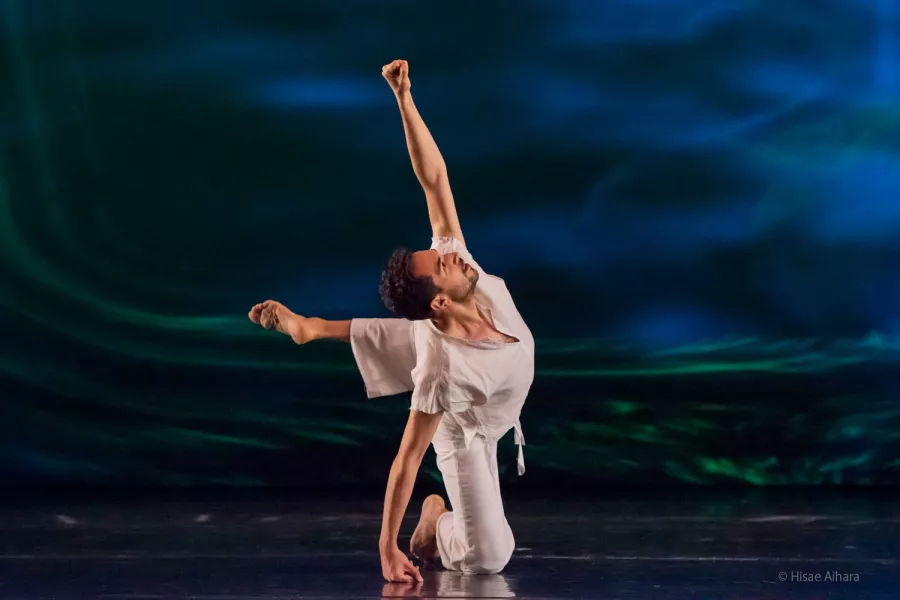A dancer dressed in all white, strikes a pose on the floor, with one knee planted on the ground, and the other raised off the ground, in a half-bent position. The backdrop is a blend of dark blue and neon green.