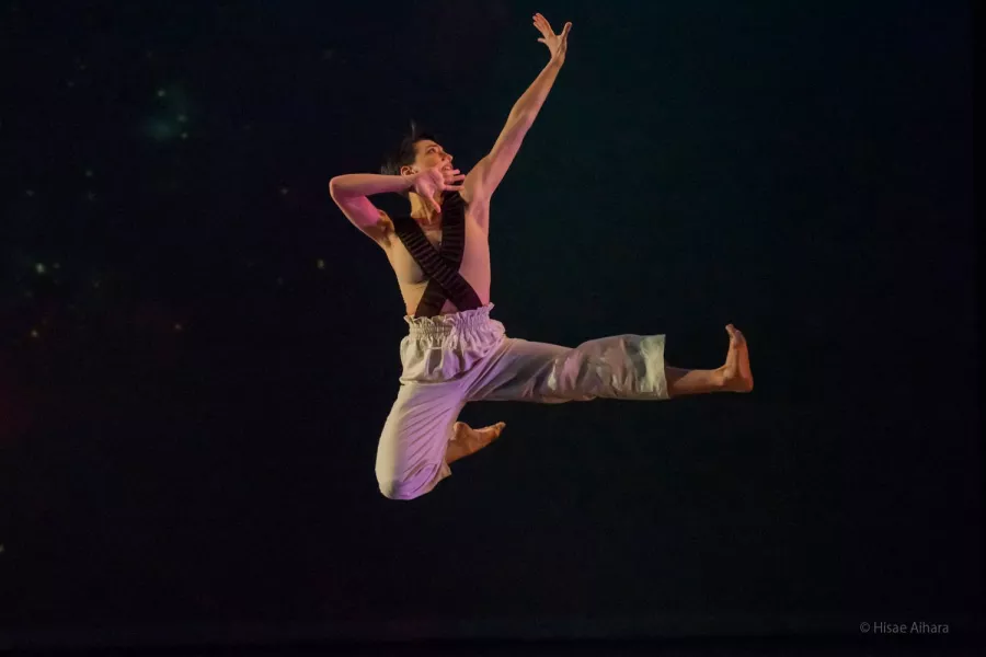 A dancer wearing pants and suspenders, jumping and striking a pose in the air: one leg horizontally fully extended, the other bent from the knee up, with their foot tucked near their lower back.