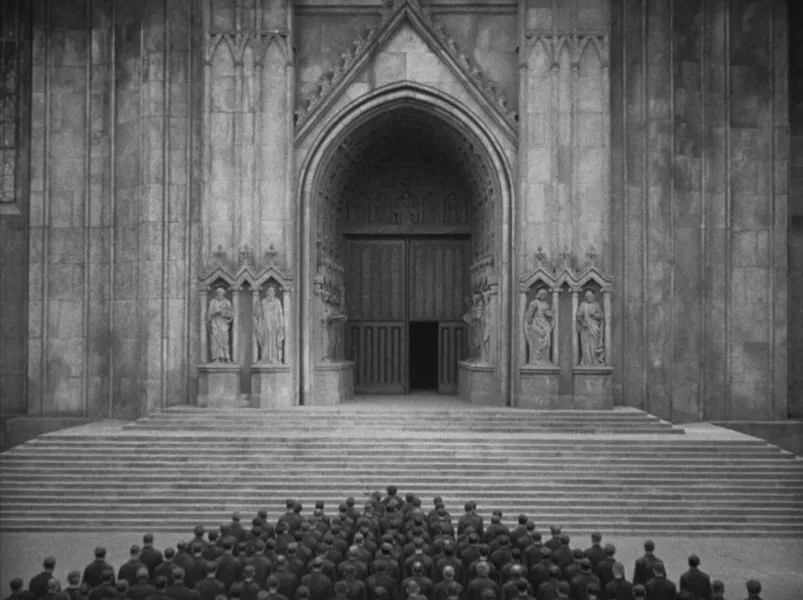 A large building with many stairs leading up to an arched opening in the wall. There are two statues of people on each side of the opening. A crowd of people are at the bottom of the stairs.