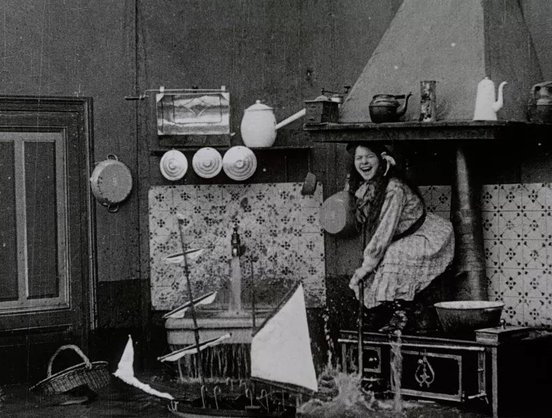A person is crouching on a raised surface laughing. There is a running sink to their right and the floor appears to be flooded. Behind the person is a white and black patterned wall.