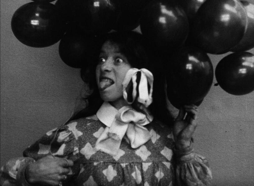 A young person looking to their right with wide eyes sticking their tongue out. They are holding balloons and have bows in their hair.