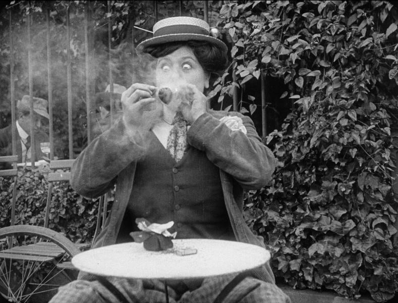 A person is smoking a cigar with wide eyes sitting at a table. They are wearing a hat and suit. Behind them are leaves.