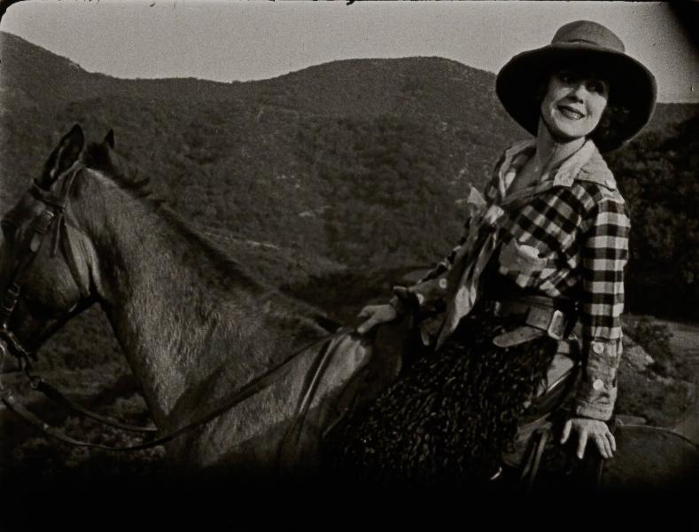 A black and white photo shows a person wearing plaid and a large hat sitting on a horse with mountains in the distance.