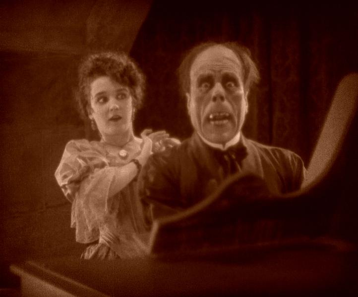 A sepia hued animation shows 2 ghosts, one with sparce, dark hair and protruding teeth wearing a tuxedo, the other in a dress with curly, brown hair, and a surprised expression, positioned behind the other. 