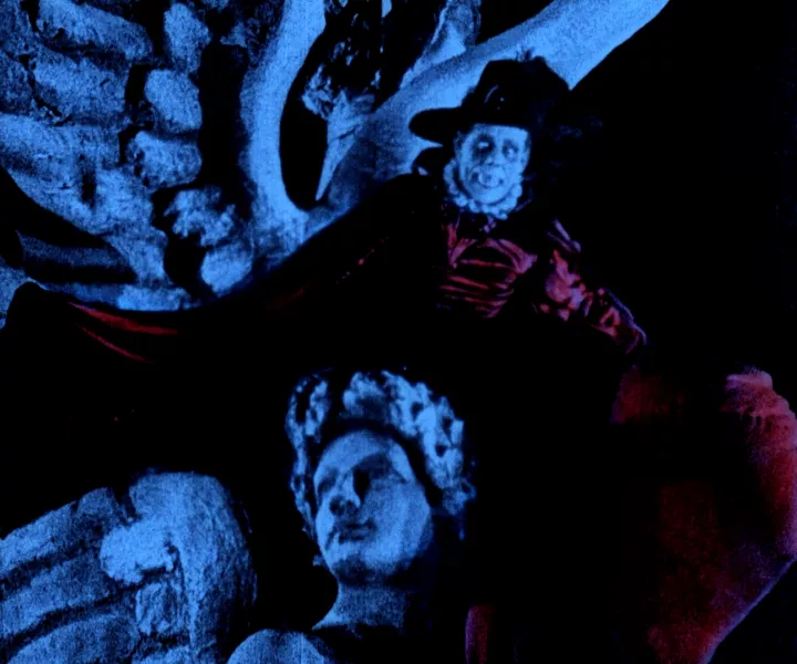 A blue hued photo shows a pale skinned character with dark eyes, a black hat, wearing a black and red cape, positioned in a hoovering stance over an angelic concrete statue.  