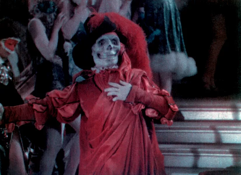 A skeleton dressed in a red dress with overlaying fabrics and ablack hat with a red feather boa attached, holds its hand over its chest with a shocked expression on its face. 