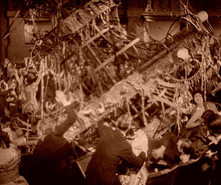 A sepia hued photo shows a large chandelier falling on top of a group of party-goers dressed in formal attire. 