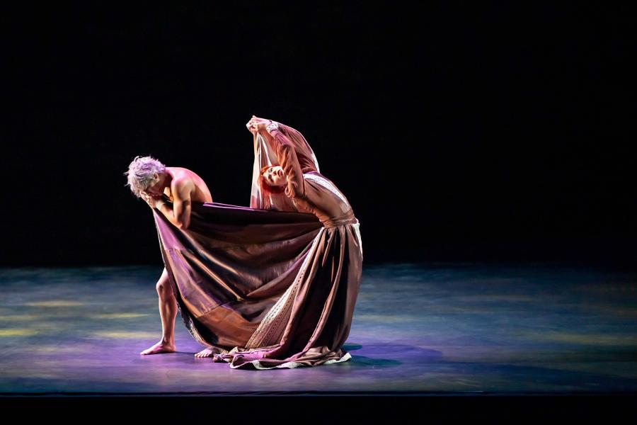 One dancer wears a long brown dress and is arching their back with their arms above their head. The dancer on the left is hiding their body behind their dress.