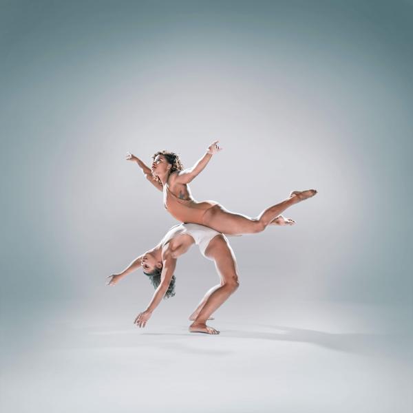 A dancer wearing white is standing arching their back with a dancer wearing beige balanced on their stomach.
