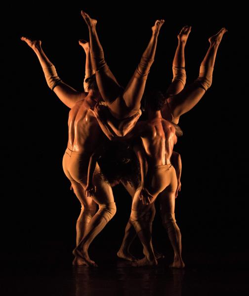 On a dimly lit stage, three dancers are held upside down in a circle while three other dancers hold them.