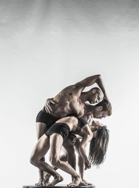 Three dancers are intertwined with two holding a third dancer who is arching their back. They are all wearing black shorts and are in front of a white and grey background.
