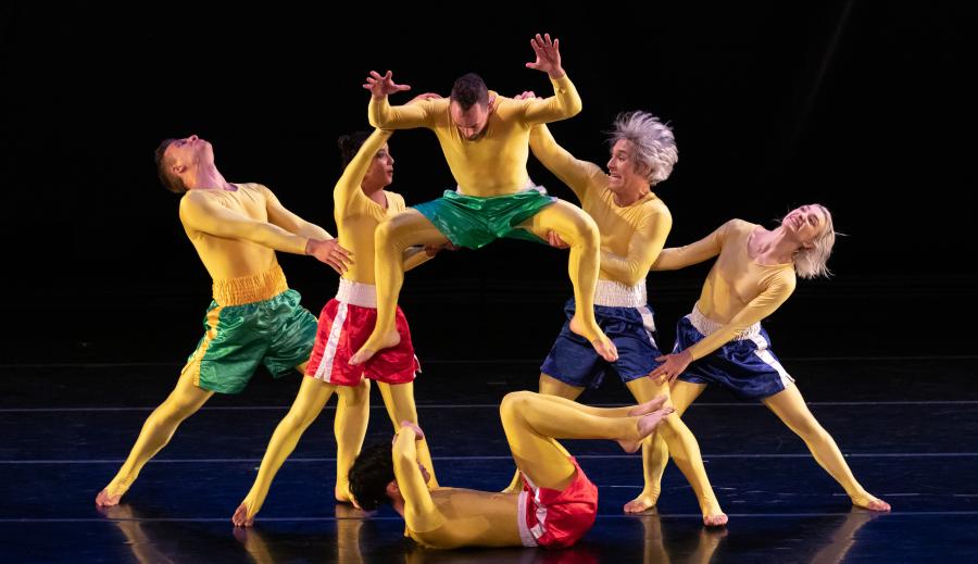 In Front of a black background, six dancers wearing yellow bodysuits and blue, green, and red shorts hold each other in various positions.