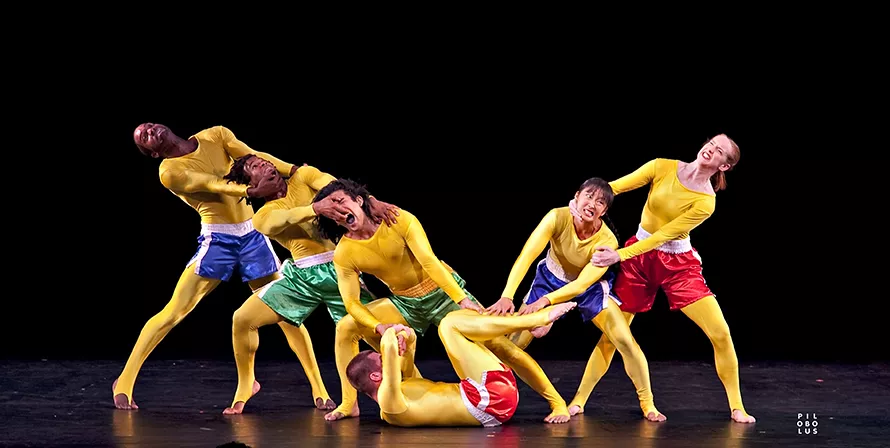 One dancer lays on their back with three on the left and two on the right. All of the dancers are pulling at each other to form a V shape and are wearing yellow bodysuits. 