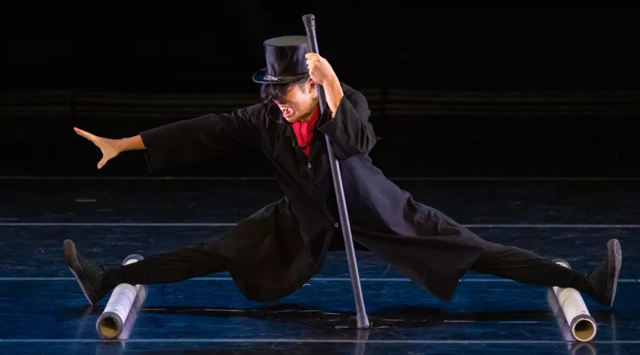 A person's legs are on two cylinders completely spread apart and nearly sitting on the ground. They are wearing a black hat and holding a cane with their left hand. The person is looking to their right.