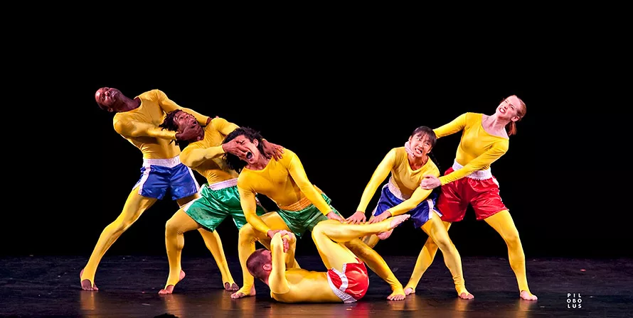 One dancer lays on their back with three on the left and two on the right. All of the dancers are pulling at each other to form a V shape and are wearing yellow bodysuits. 