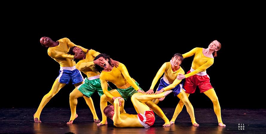 One dancer lays on their back with three on the left and two on the right. All of the dancers are pulling at each other to form a V shape and are wearing yellow bodysuits. 