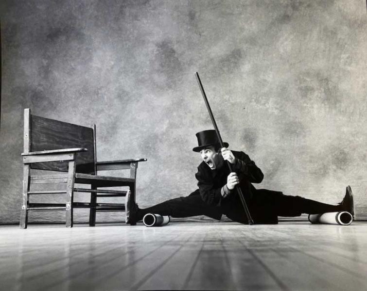A person's legs are on two cylinders completely spread apart and nearly sitting on the ground. They are wearing a black hat and holding a cane. The person is looking to their right at a chair.