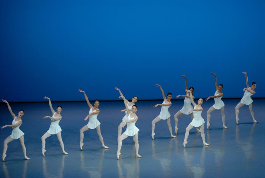 Ten dancers stand with a straight left leg and right leg forming a 90 degree angle at the knee. Their arms are straight with the right arm lifted above the left. Their dresses are white and the background is blue.