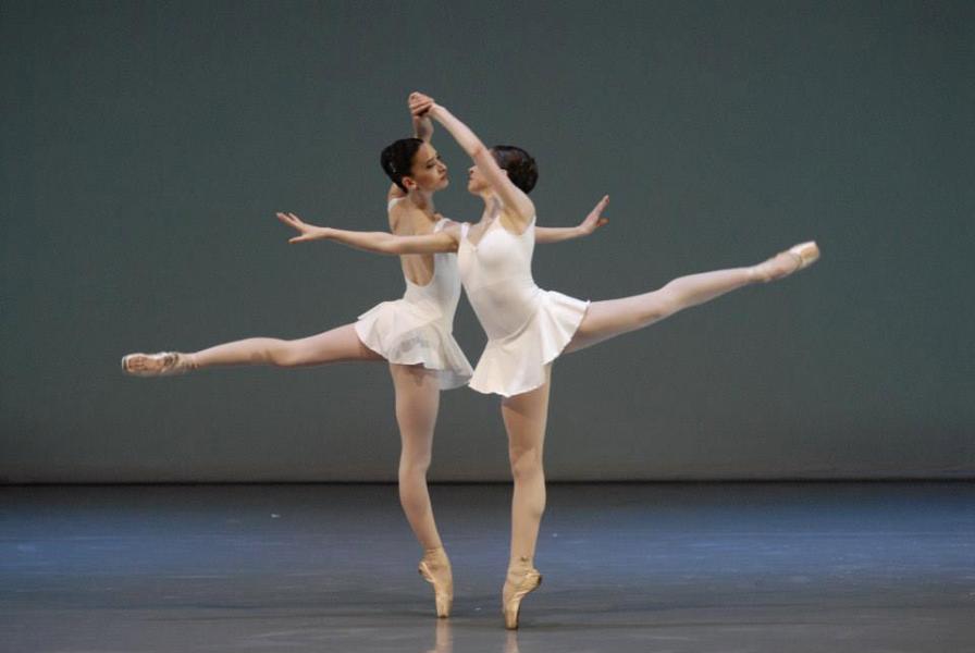 Two dancers stand side by side holding each other's left hand above their head. One leg is extended behind them. Their dresses are short and white. The background is grey and blue. 