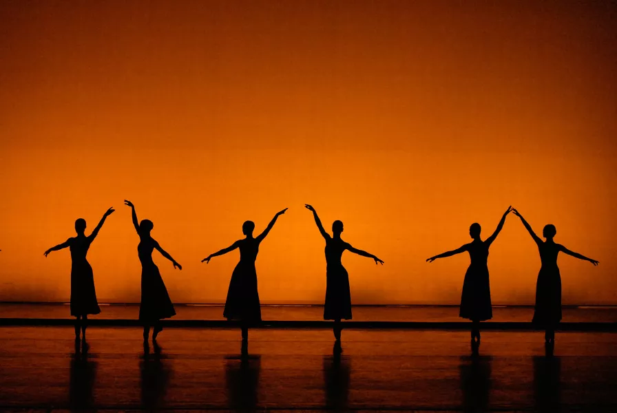 Three dancers stand with their right arm at an angle and three with their left in front of an amber background.