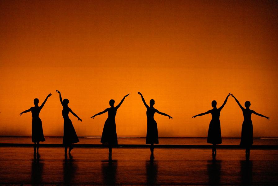 Three dancers stand with their right arm at an angle and three with their left in front of an amber background.