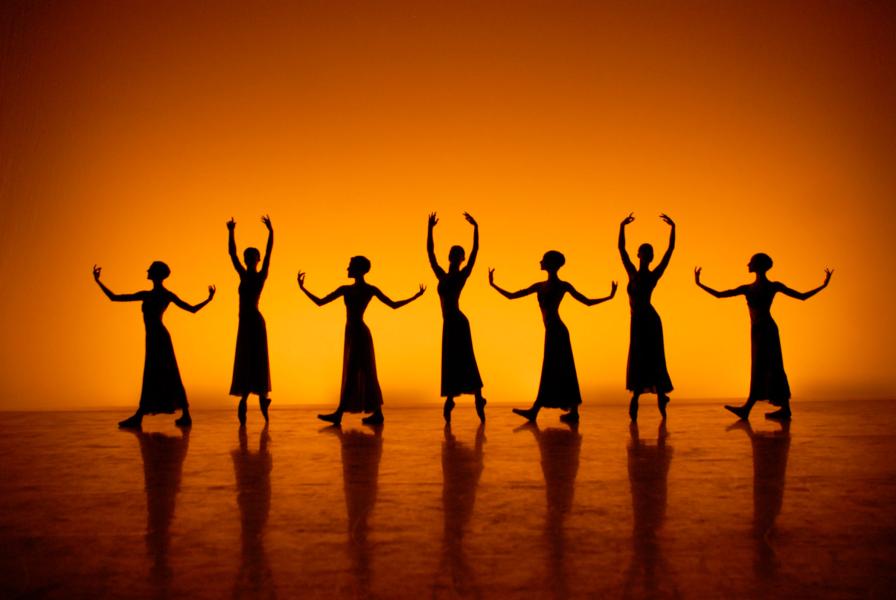 A stage is lit with a golden yellow hue, revealing the shadows of seven dancers, four of which hold their arms in a position parallel with the floor, three of which hold their arms above their heads.