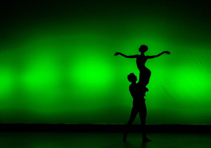 A silhouette of a dancer carrying another dancer by their legs appears in front of a green background. The lifted dancer's arms are a straight line.
