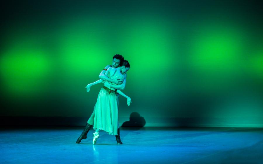 The stage is lit with a lime green hue. One dancer hugs another whose arms are extended to the side.