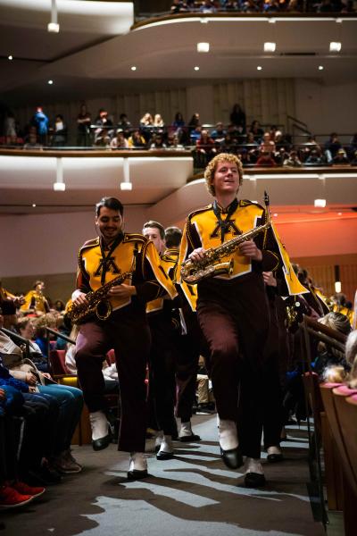 Two UMN band members lead a group behind them holding gold saxophones. They are wearing yellow tops and maroon bottoms. People are seated on both sides of them.