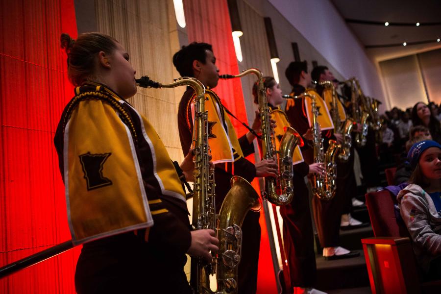 A row of UMN marching band members, looking away from the camera, play a saxophone in front of a wall that is lit with maroon and gold lights. 