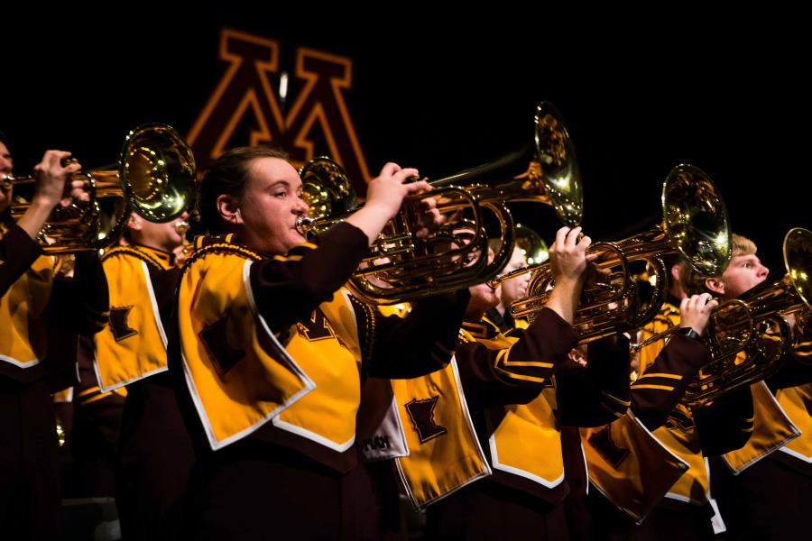 Two horizontal lines of UMN Marching Band members performing on stage, wearing maroon and gold, holding their trumpets into the air.