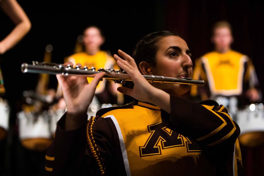 A UMN marching band member dressed in maroon and gold, playing the flute on stage. The camera focus is on the one player, while two drum players are blurred in the background.
