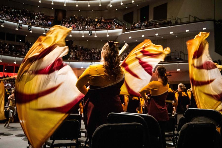 The backs of a three UMN Marching Band members performing on stage, wearing maroon and gold, waving large flags into the air.