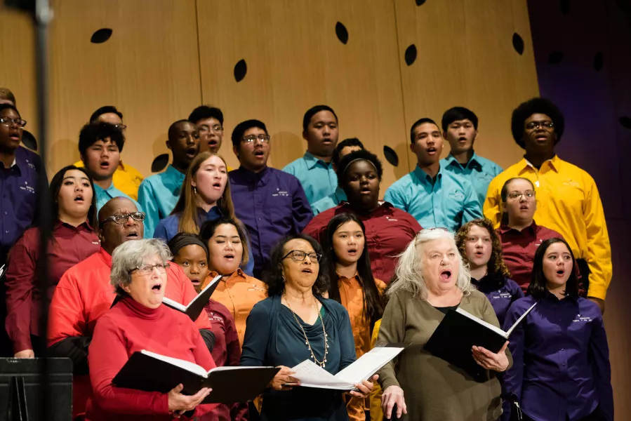 A large choir group wearing an assortment of colors, sing together, lined up in vertical rows of increasing height.