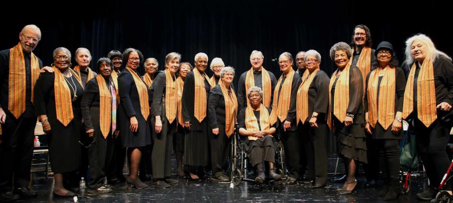 A group of choir performers, wear all black with yellow sashes around their neck, smiling for a group photo.