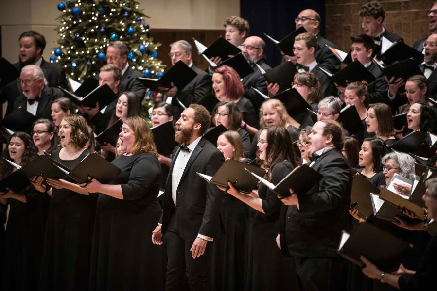 A large group of varying genders and ages all dressed in black and white, stand on elevated rows, singing in choir, with a green, lit Christmas tree in the background.